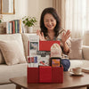 A smiling woman sits on a sofa behind a wooden coffee table featuring a red gift hamper filled with gourmet snacks, tea, and treats.